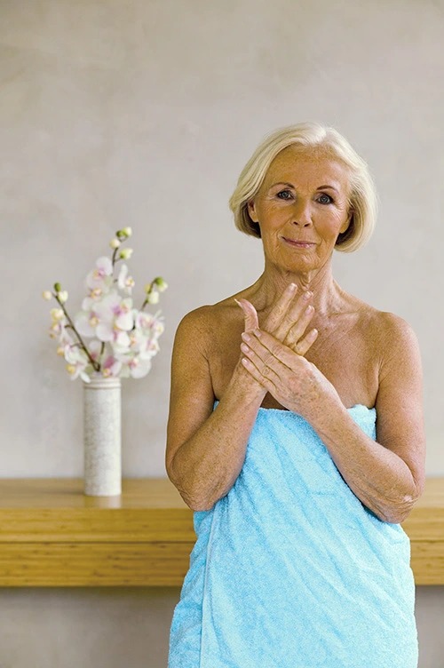 Senior woman wrapped in a light blue towel standing by a wooden shelf with white orchids looking relaxed